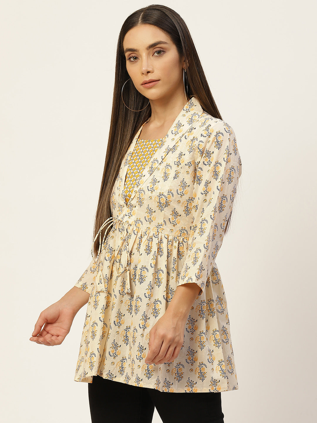 A woman modeling a cream-colored, three-quarter sleeve tunic top with a blue and yellow floral print, featuring a tie waist and a gold patterned inner neckline, set against a plain light background.