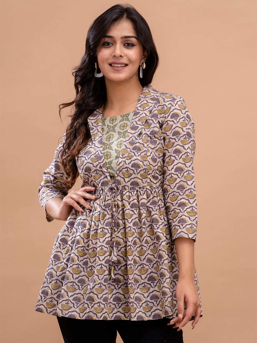 A woman modeling a three-quarter sleeve printed tunic top with a drawstring waist, featuring a brown and beige geometric pattern with a green embroidered neckline, set against a solid beige background.