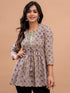 A woman modeling a three-quarter sleeve printed tunic top with a drawstring waist, featuring a brown and beige geometric pattern with a green embroidered neckline, set against a solid beige background.